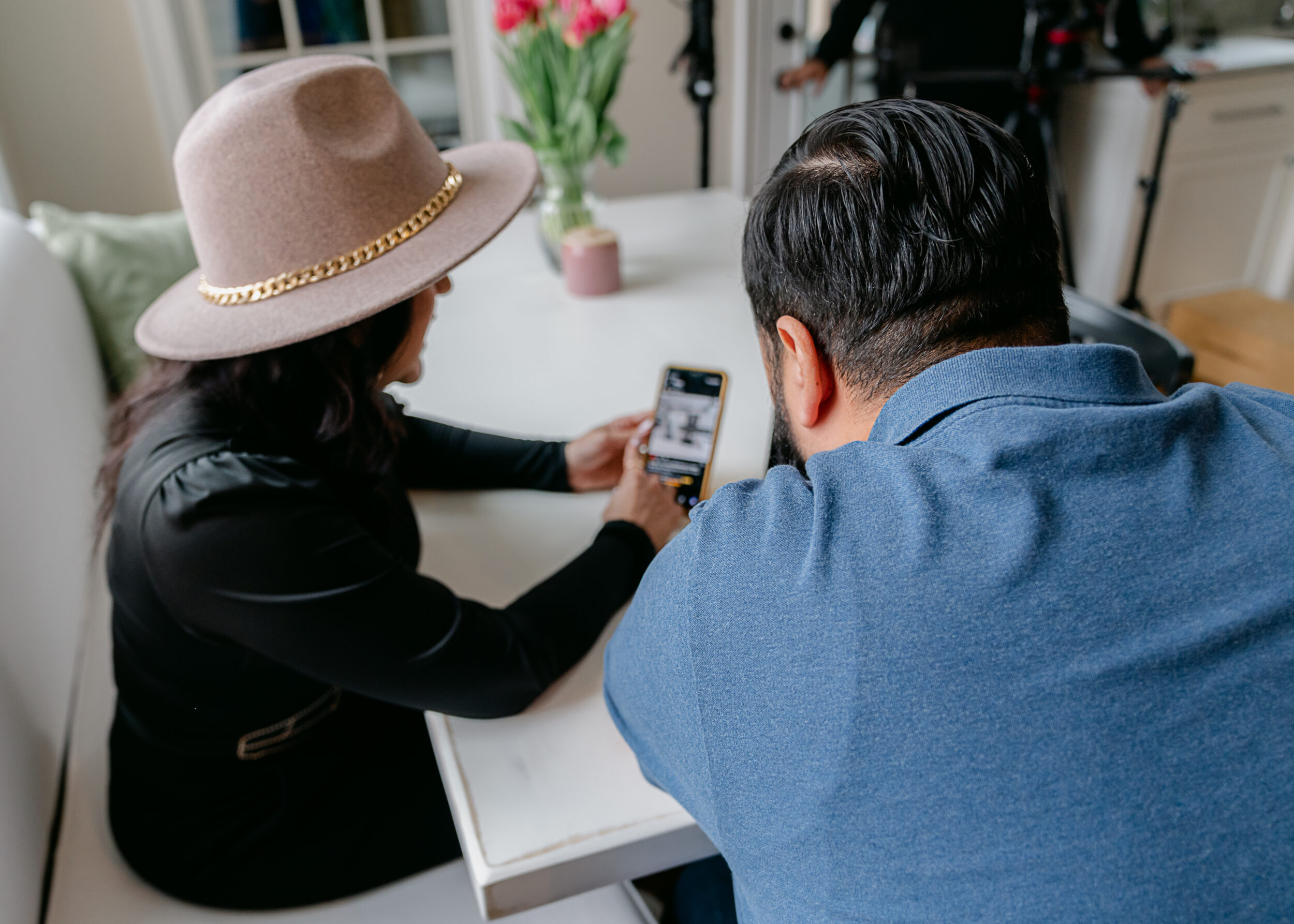 Girl with hat showing man in blue polo her phone at a dining room table